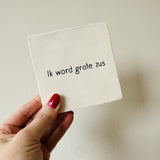 Wit vierkant tegeltje met de zwarte tekst 'Ik word grote zus', vastgehouden in een hand tegen een lichte muur.