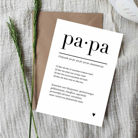 Witte ansichtkaart Papa met zwarte tekst en een klein hartje, op een bruine envelop naast groene takjes.
