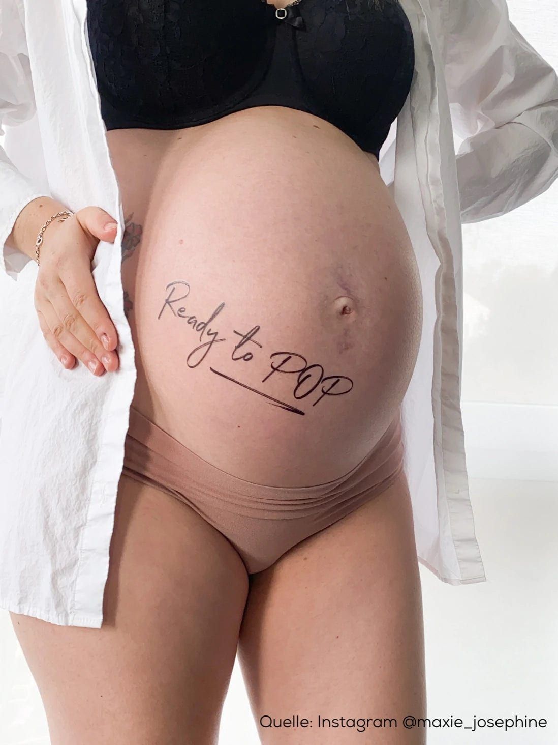 Zwarte plaktattoo 'Ready to Pop' op een zwangere buik, met een open wit overhemd en een bruine slip.