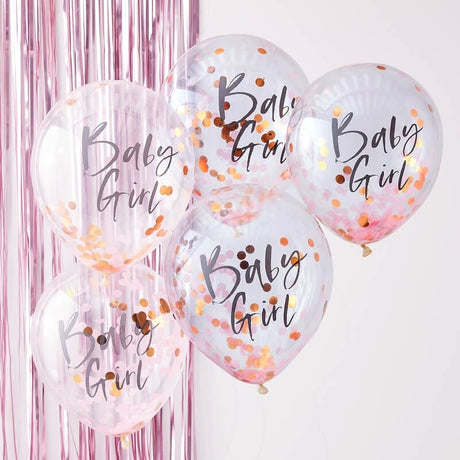 Vijf transparante ballonnen met 'Baby Girl' en roze en goudkleurige confetti voor een roze franjesachtergrond.