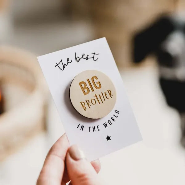 Ronde houten Big Brother badge op een witte kaart met de tekst 'the best in the world', vastgehouden in een hand.