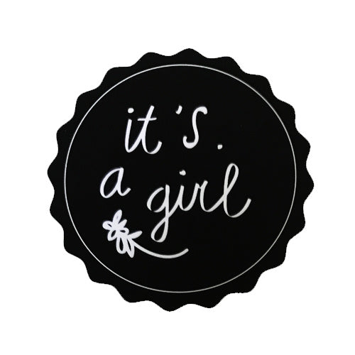 Zwarte ronde sticker met geschulpte rand, witte tekst ‘it's a girl’ en een klein bloemetje.