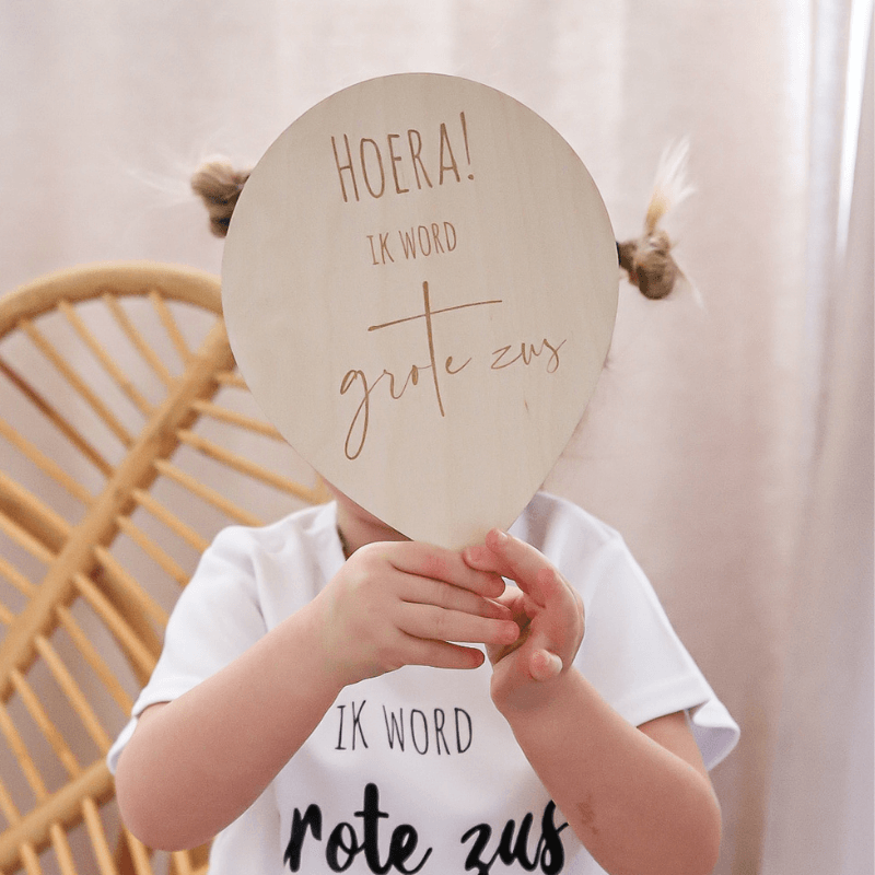 Kind in wit shirt houdt een houten ballon met de tekst 'Hoera ik word grote zus' voor het gezicht.