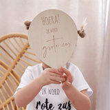 Kind in wit shirt houdt een houten ballon met de tekst 'Hoera ik word grote zus' voor het gezicht.