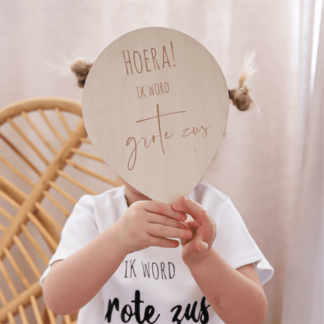Kind in wit shirt houdt een houten ballon met de tekst 'Hoera ik word grote zus' voor het gezicht.