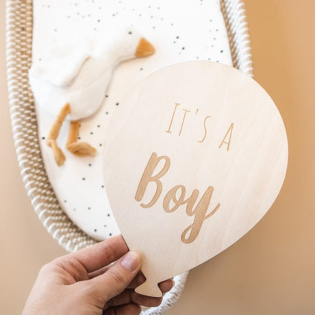 Hand houdt een houten ballon met opdruk ‘It's a boy’ vast voor een rieten mand met een witte vogelknuffel.