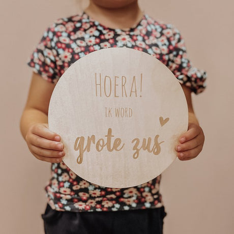 Kind houdt een rond houten bord vast met de tekst 'Hoera! Ik word grote zus' en een hartje.