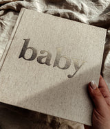 Hand houdt het naturelkleurige baby-invulboek met goudkleurige titel vast boven een geplooide stof.