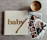 Linnen baby-invulboek met naturelkleurige kaft en gouden tekst 'baby', naast babyfoto's en een potlood.