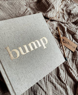 Bump zwangerschapsinvulboek op een geplooide doek, met goudkleurige titel en een potlood ernaast.