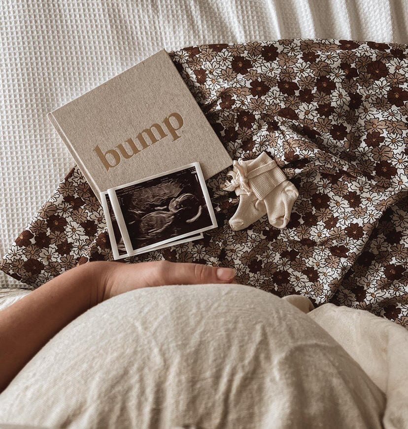 Bump zwangerschapsinvulboek naast echofoto's en babysokjes op een bloemendoek, met een zwangere buik op de voorgrond.