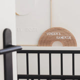 Houten regenboogletterbord met witte letters op een plank achter een zwart babybed.