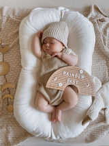 Slapende baby in een wit babynest met het houten regenboogletterbord 'rainbow baby' op de buik.