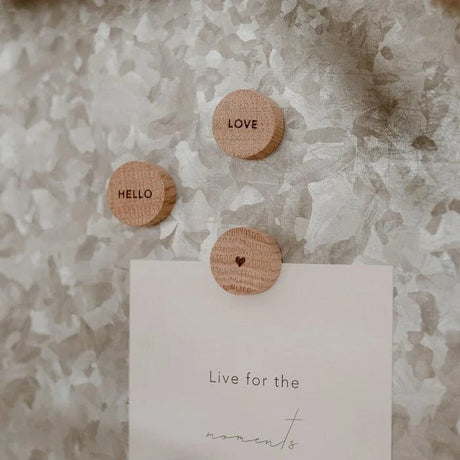 Drie ronde eikenhouten Hello Love-magneten met ‘Hello’, ‘Love’ en een hartje, waarvan één een kaart vasthoudt.