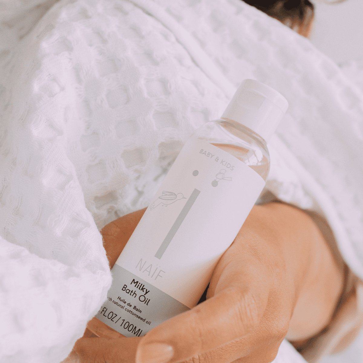 Hand houdt een fles Naïf Milky Badolie vast tegen een witte badjas met reliëf
