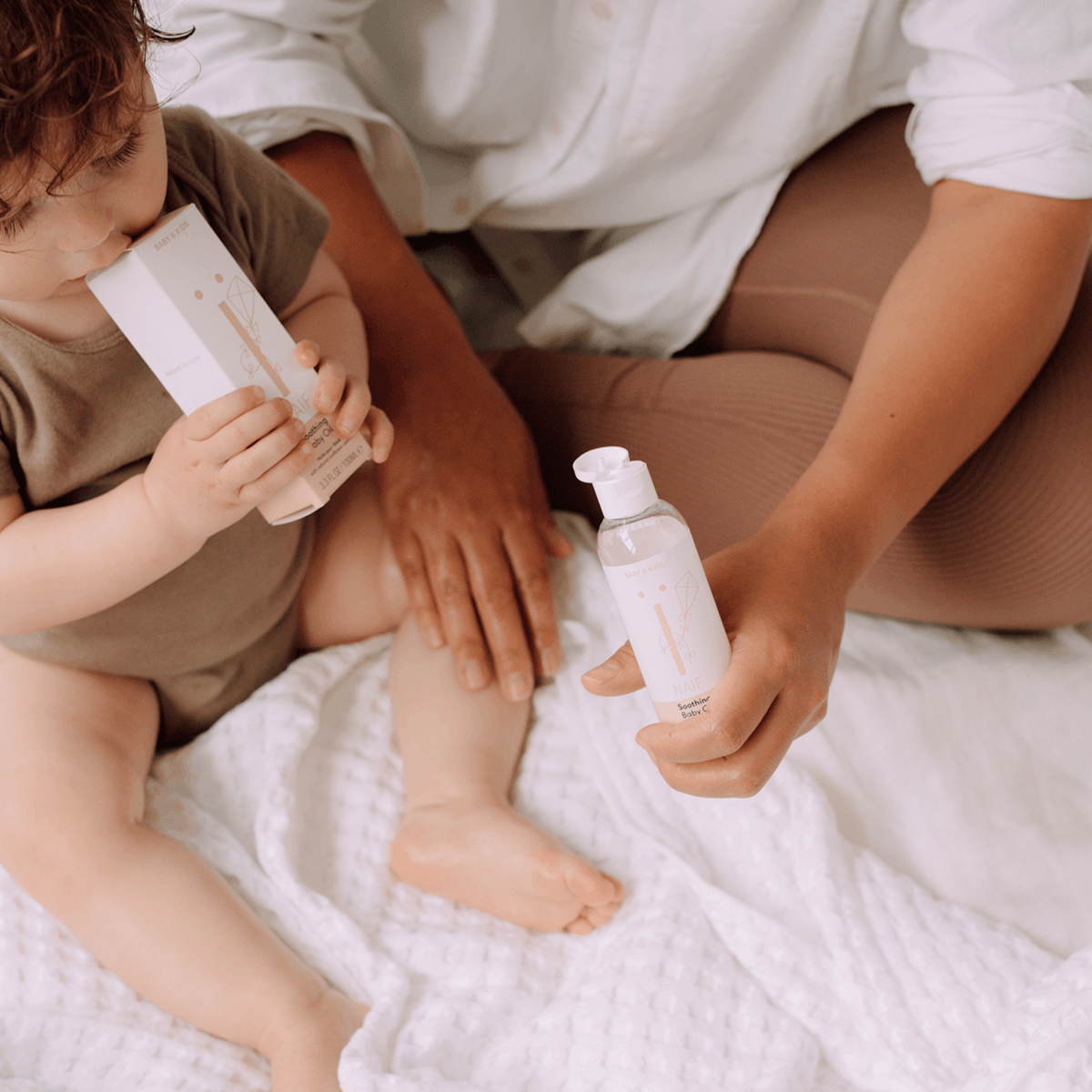 Een volwassene houdt Naïf Verzachtende Babyolie vast en masseert een babybeen terwijl de baby de verpakking vasthoudt.