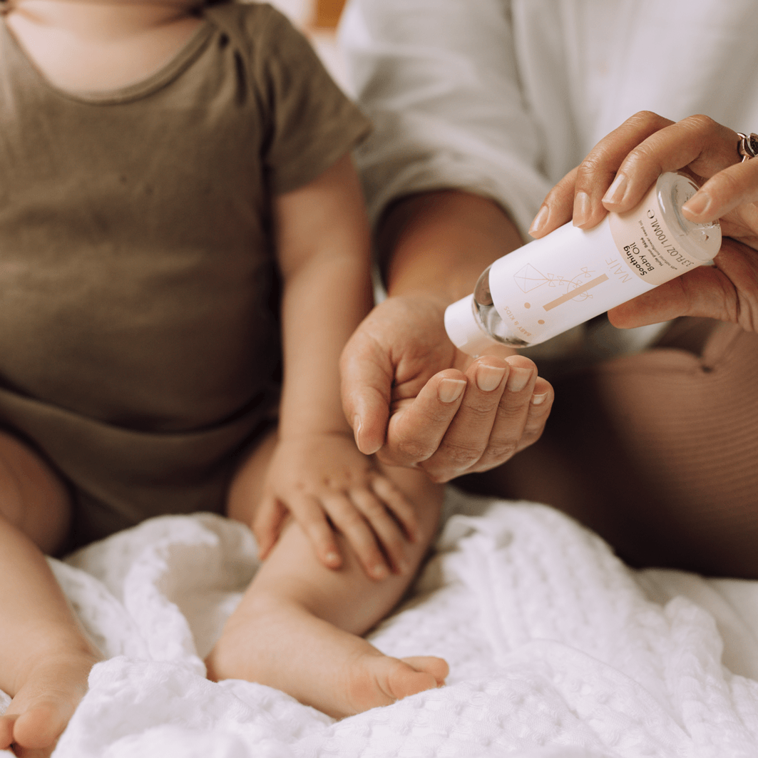 Een volwassene schenkt Naïf Verzachtende Babyolie in de hand naast een baby op een witte deken.