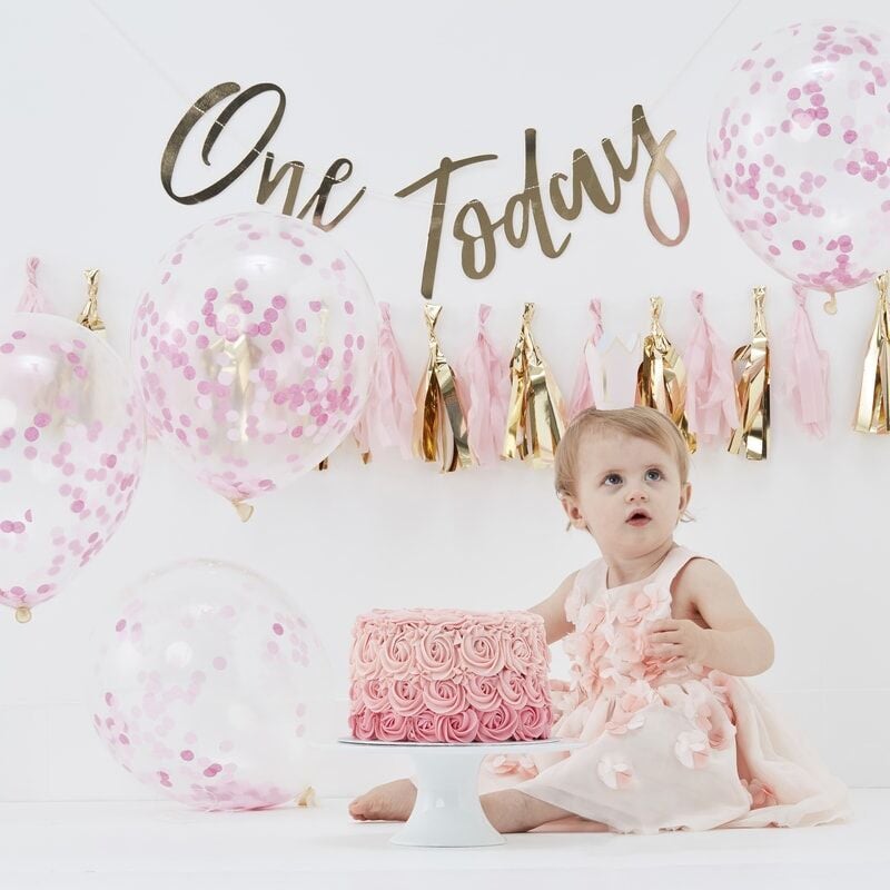 Roze cake smash kit met confettiballonnen, roze en gouden slingers en een baby naast een roze taart.