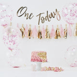 Roze cake smash kit met confettiballonnen, kwastjesslinger en gouden 'One Today'-slinger achter een aangesneden taart.