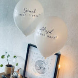 Witte troostballonnen 'Straal maar liefje' en 'Altijd in ons hart' boven een ingelijste prent naast een kaars.