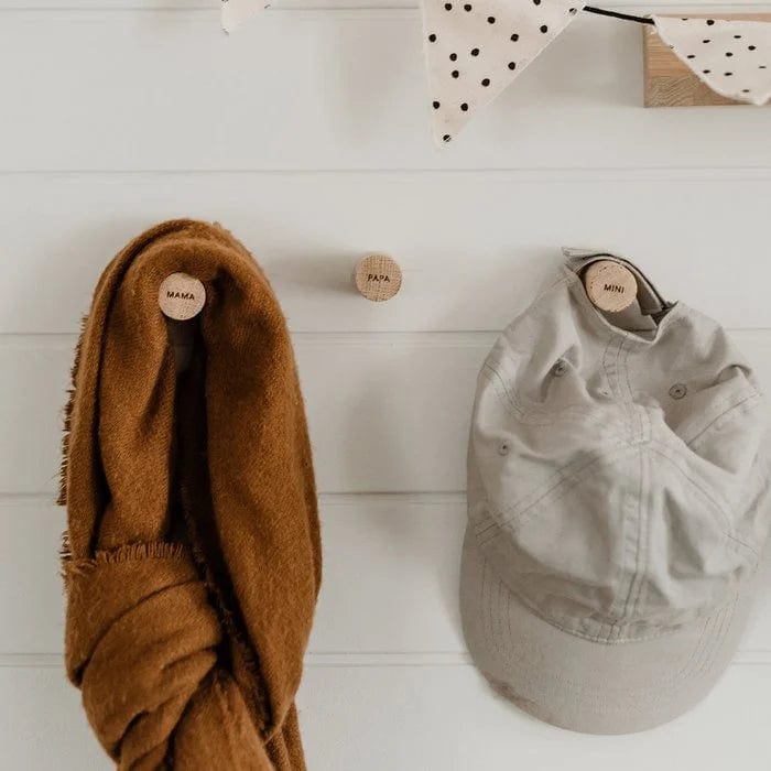 Houten wandhaken Mama, Papa en Mini aan een witte wand, met een bruine sjaal en lichtgrijze pet.
